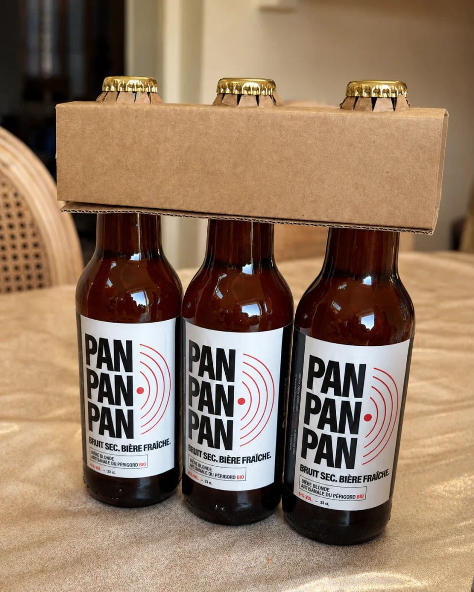 Photographie de trois bouteilles PAN PAN PAN étiquetées et réunies sous une poignée en carton, posées sur une table.