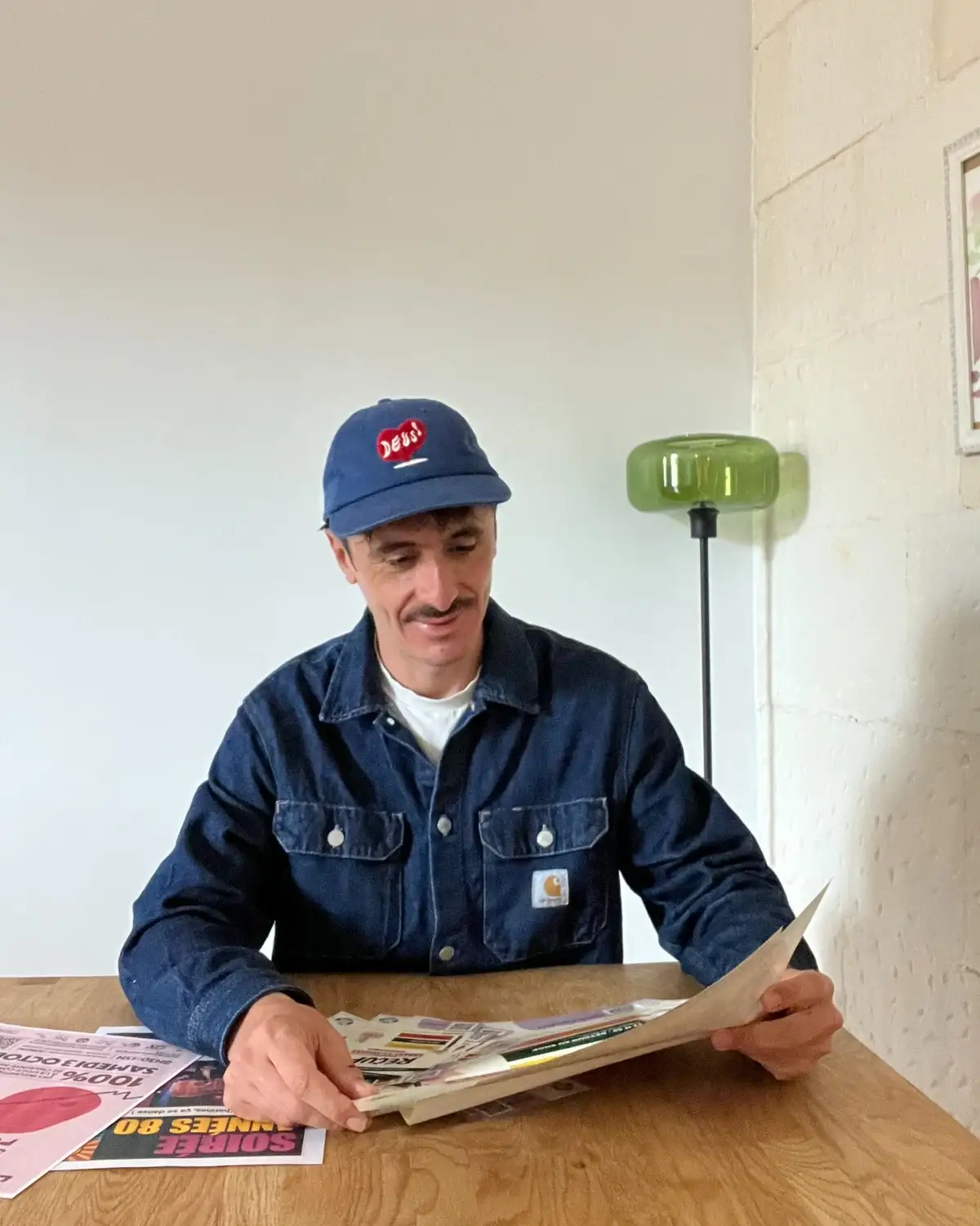 Cédric Chamoulaud, casquette bleue, examinant des supports imprimés et des maquettes de communication à son bureau.